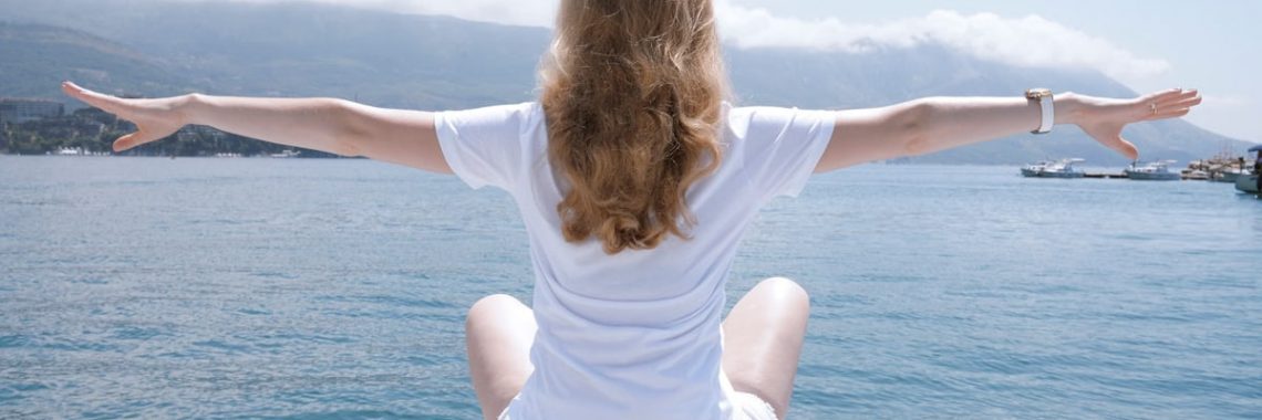 Woman sitting with open arms by the water, embracing nature and serenity.